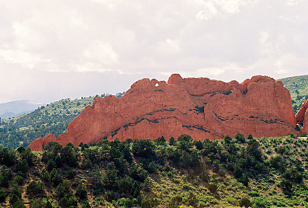 [A large red cliff-like rock with a section at the top with a hole in the middle. There is some greenery growing at lower levels along ledges, but it's a nearlly all reddish rock.]